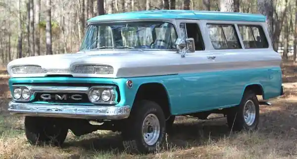Классический бирюзово белый автомобиль GMC Suburban стоит на фоне деревьев.