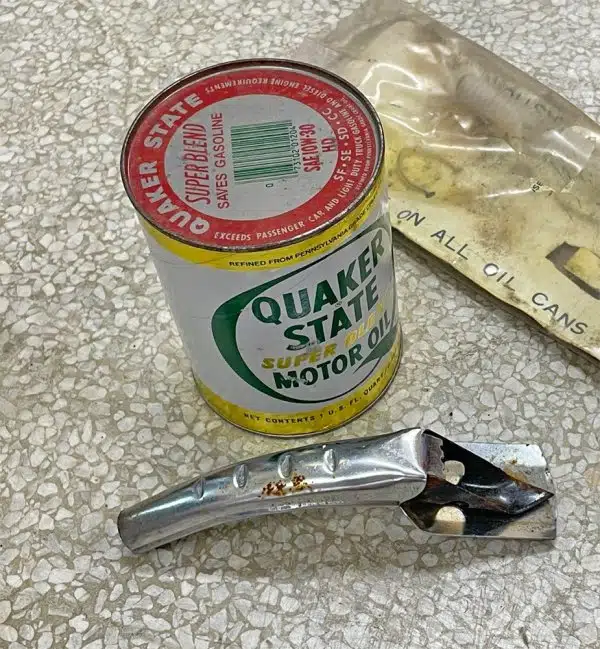 На фактурной поверхности лежат винтажная банка с моторным маслом Quaker State и старый металлический инструмент для ее открывания и заливки.