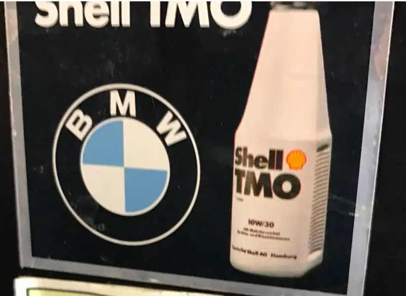 Классический круглый знак с белым и синим орнаментом и буквами BMW на темном фоне представлен вместе с упаковкой моторного масла Shell TMO.