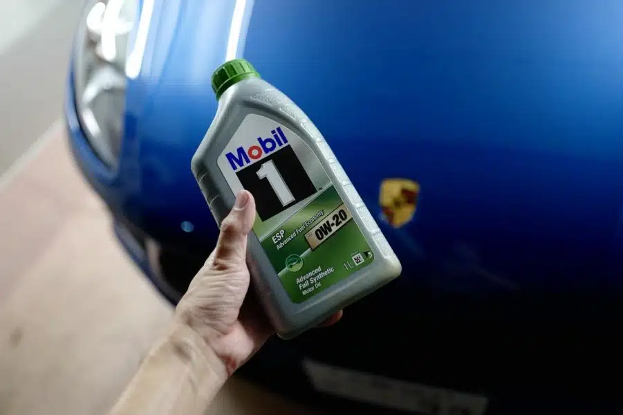 Рука держит моторное масло Mobil 1 на фоне синего автомобиля Porsche показывая его готовность к техническому обслуживанию