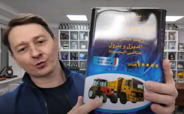 Мужчина на фоне стеллажей с моторным маслом демонстрирует большую металлическую банку с рисунками сельскохозяйственной техники и грузовика.