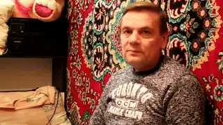 Мужчина на фоне ковра и мягких игрушек смотрит в камеру в вязаном свитере.