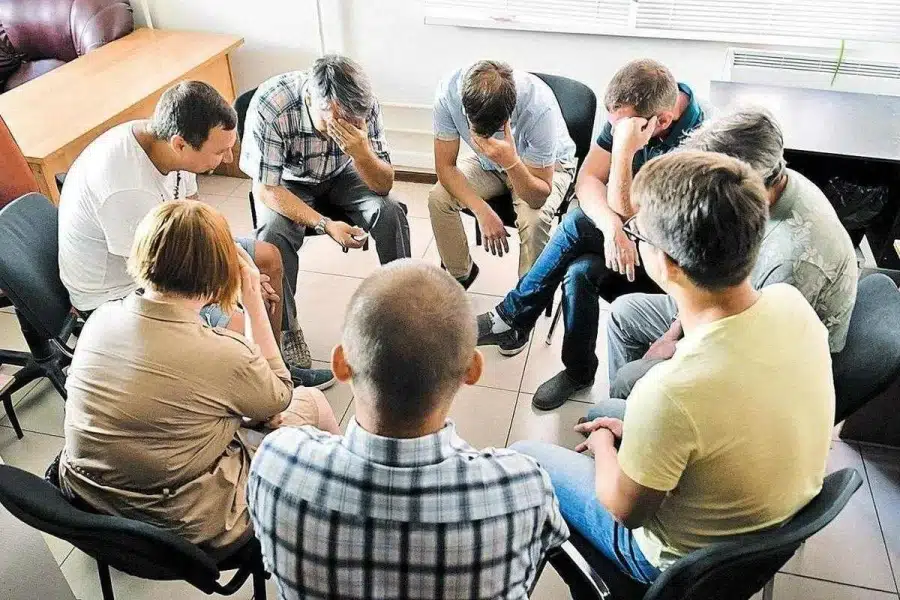 В комнате собрались восемь человек в кругу на стульях трое закрыли лица руками выражая переживания их ноги направлены к центру.