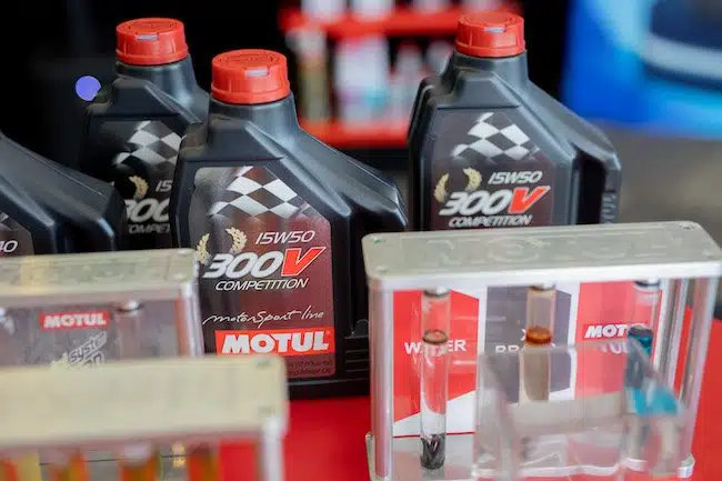 На изображении моторное масло Motul 300V Competition в черных канистрах с красными крышками и образцы жидкостей в пробирках.