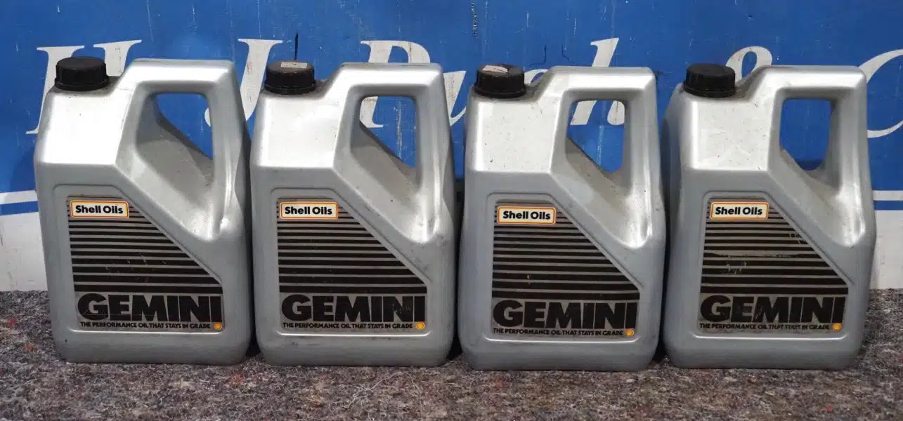 Четыре серебристые канистры с черными крышками и ярлыками Shell Oils Gemini ровным рядом расположены на поверхности перед синим фоном с белыми буквами.