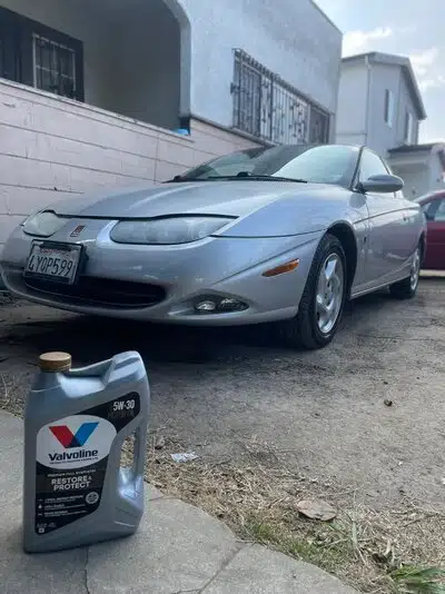 Серебристый автомобиль Saturn и моторное масло Valvoline на фоне дома в солнечный день.