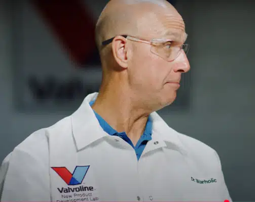 Мужчина в белом халате Valvoline и очках смотрит в сторону, вероятно, в лаборатории.