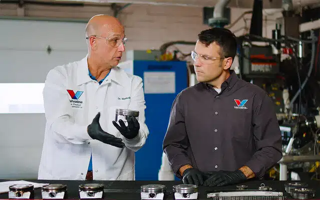 Два техника Valvoline осматривают поршни двигателя в лаборатории, обсуждая детали.