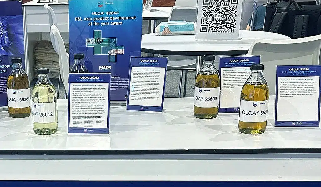 На белом столе представлены бутылки с жидкостями OLOA и рекламные материалы на выставке, демонстрирующие продукты и награды.